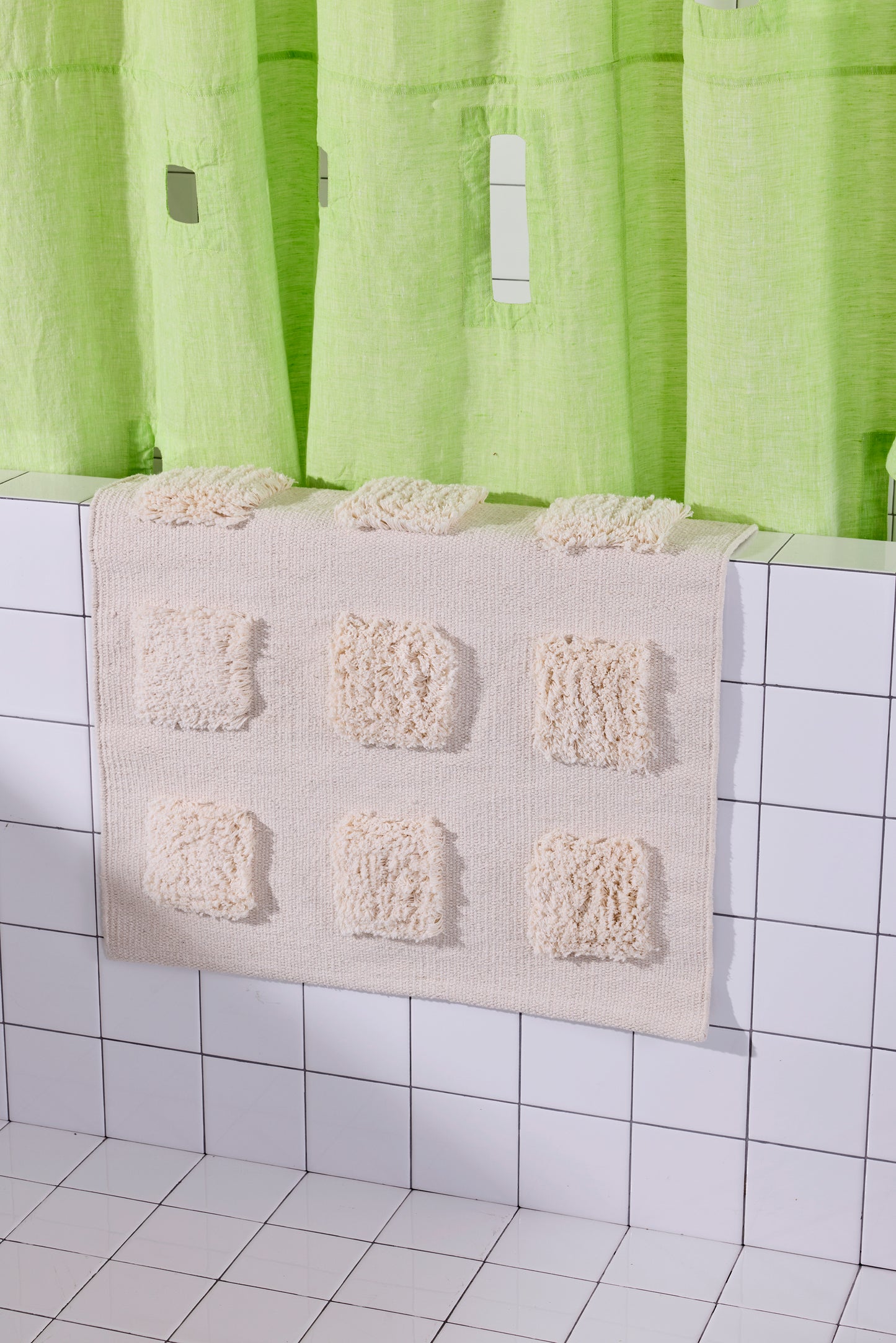 cream bath rug with raised tufted squares in natural tones, draped over a white tiled bathroom ledge beneath a lime green shower curtain.