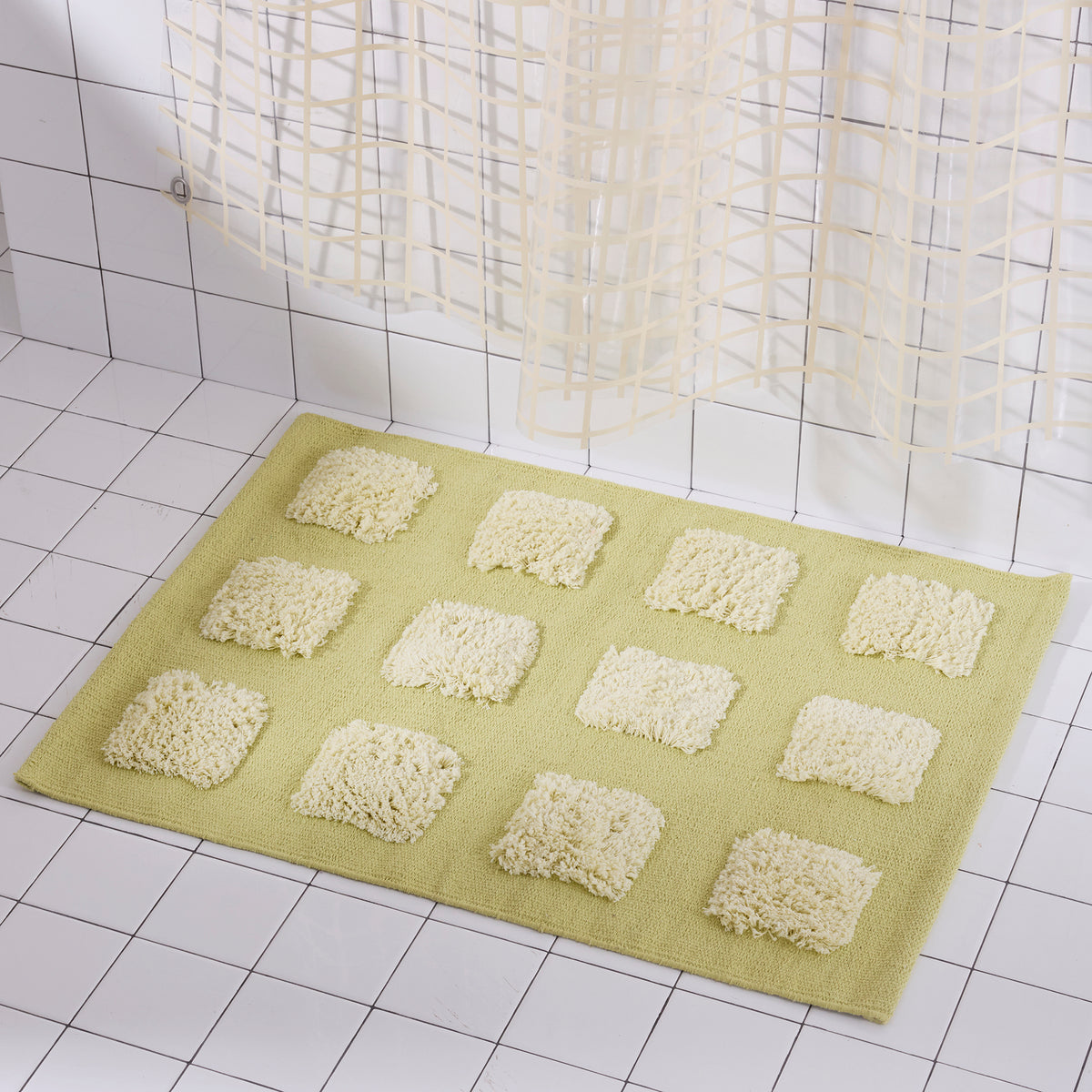 A rectangular bath mat in pale yellow-green with raised light yellow quares, shown on a white tiled bathroom floor near a shower curtain.