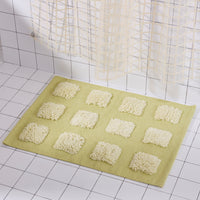 A rectangular bath mat in pale yellow-green with raised light yellow quares, shown on a white tiled bathroom floor near a shower curtain.