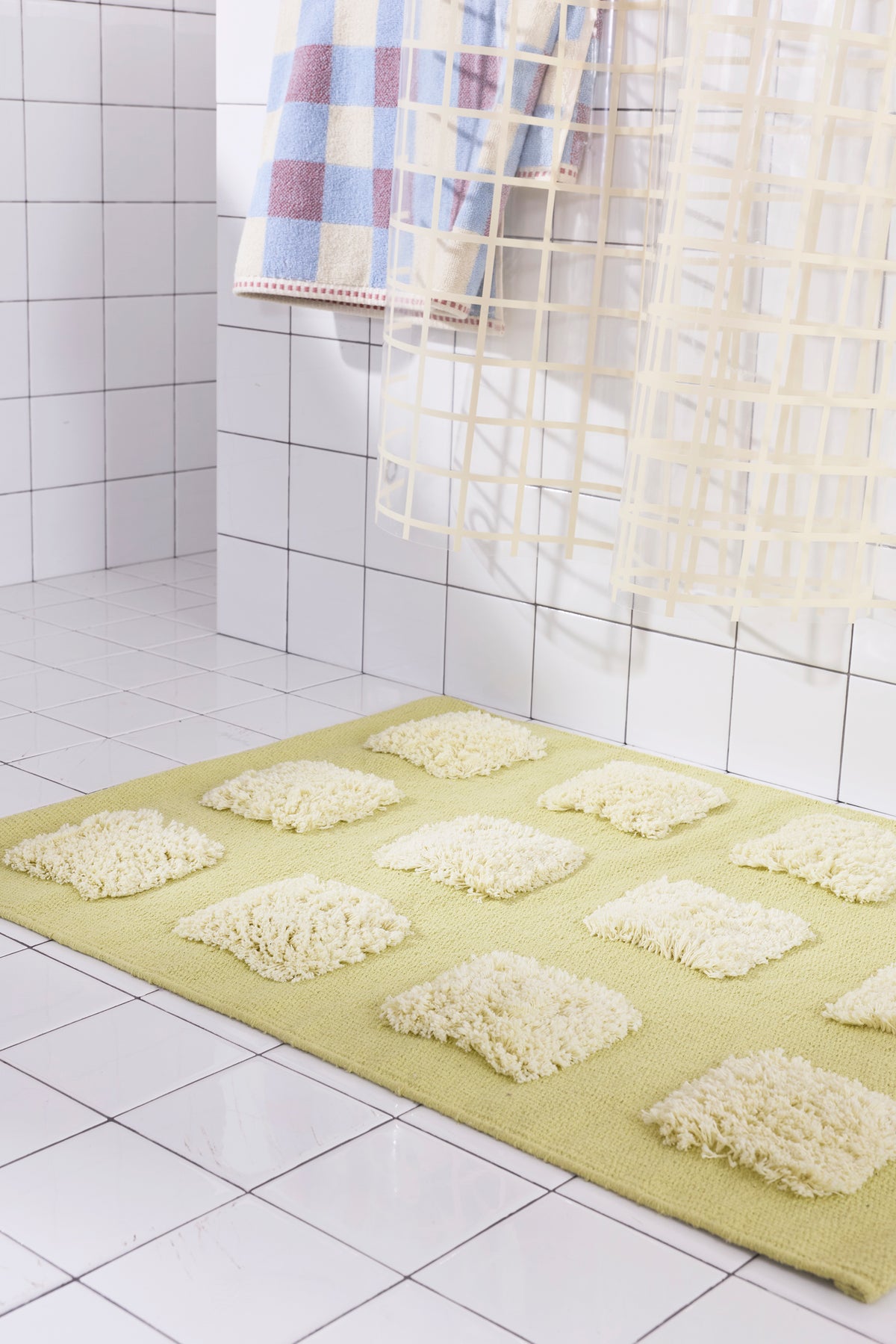 Close-up of the yellow/olive, natural bath rug showing its textured woven base and raised plush tufted squares in warm neutral shades beneath a cream shower curtain.