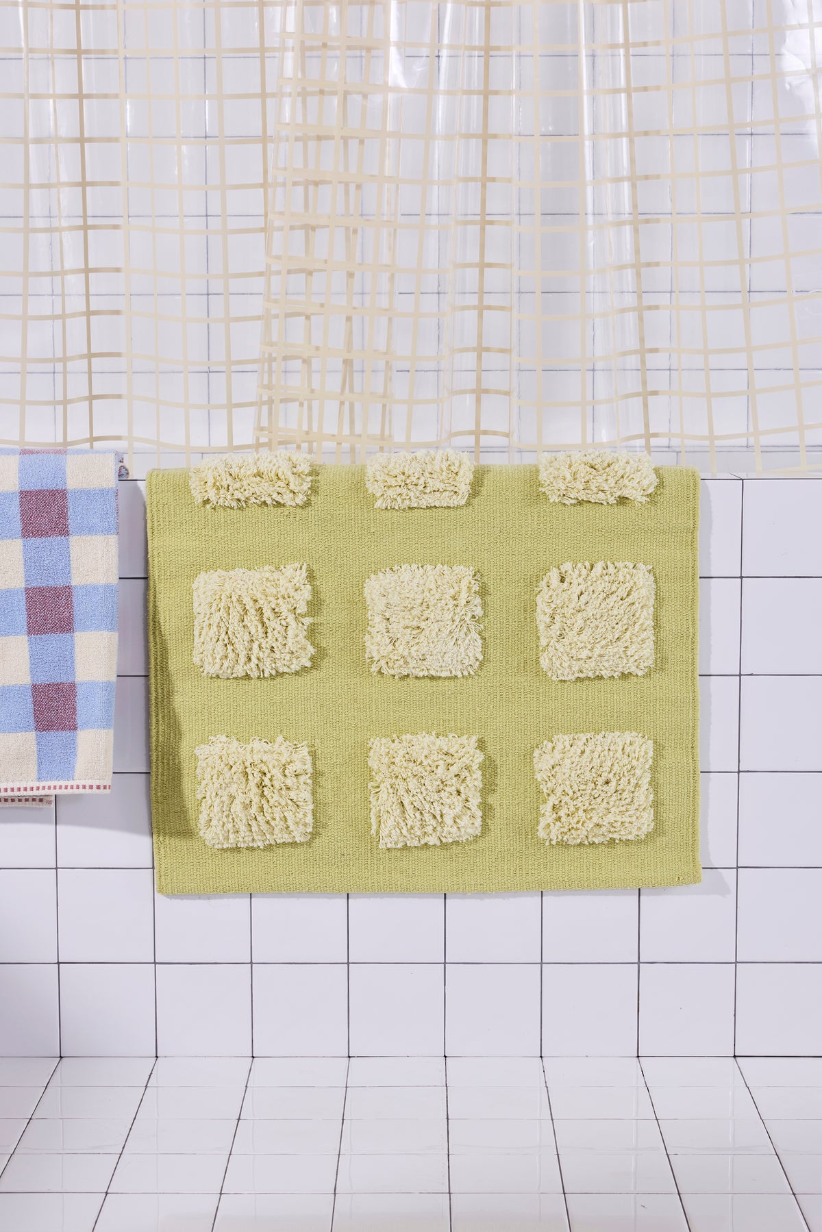yellow/olive bath rug with raised tufted squares in natural tones, draped over a white tiled bathroom ledge beneath a cream shower curtain.