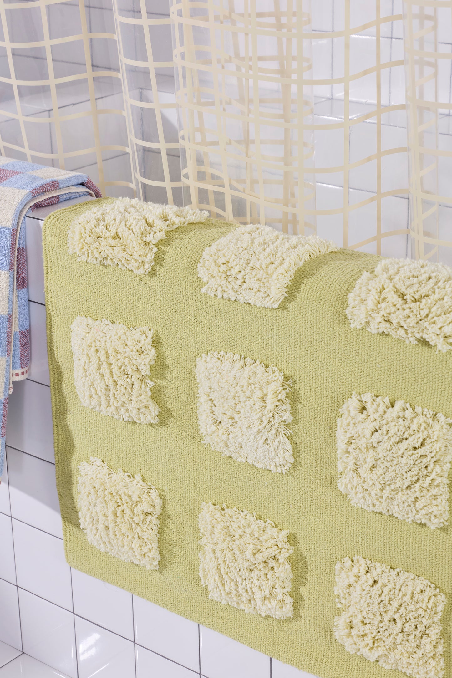 yellow/olive bath rug with raised tufted squares in natural tones, draped over a white tiled bathroom ledge beneath a cream shower curtain.