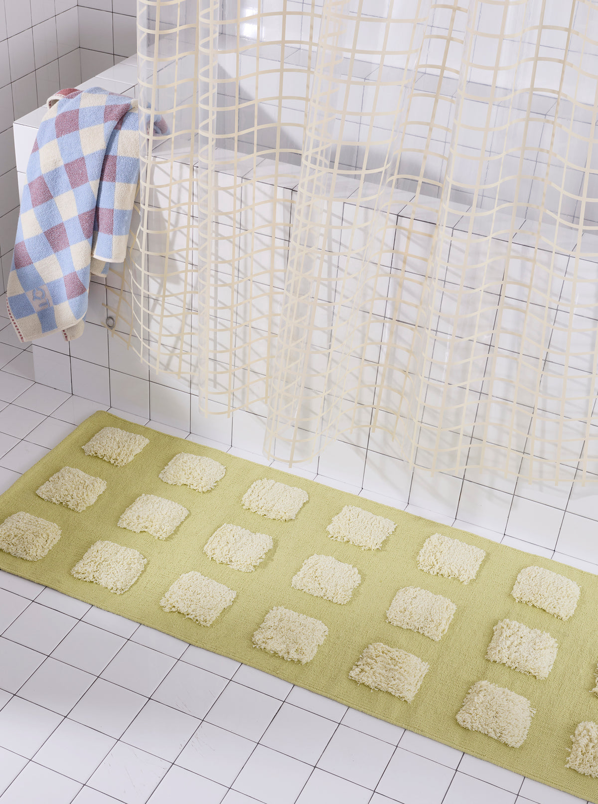 Close-up of the yellow/olive, natural bath runner showing its textured woven base and raised plush tufted squares in warm neutral shades beneath a cream shower curtain.