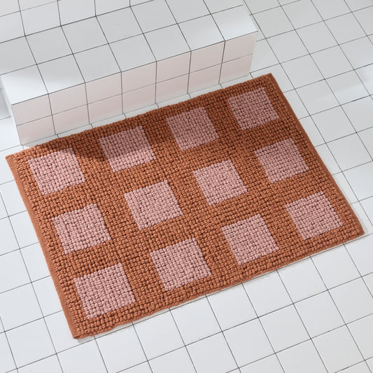 Full view of the Portal Clay bath mat in clay and blush pink geometric square grid pattern on white tile.